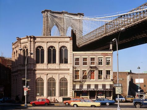 Looking north at Brooklyn Bridge, spanning East River between Brooklyn & Manhattan, New York City, New York County, NY. View of Brooklyn tower emerging behind nineteenth century commercial buildings on the corner of Front Street and Old Fulton Street (also called Cadman Plaza West).